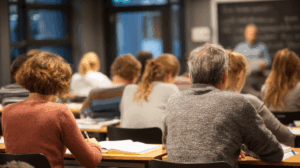 A group of adults in an evening English class, listening to the teacher and taking notes.