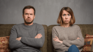 A man and a woman sitting on a couch facing opposite directions, with exaggerated annoyed expressions real people