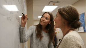 A teacher correcting a student’s sentence on a whiteboard that says “Does she likes ice cream?” with a red mark and correction.