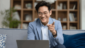 An Asian man in business casual attire speaking during a video call at home, smiling and pointing at his screen.