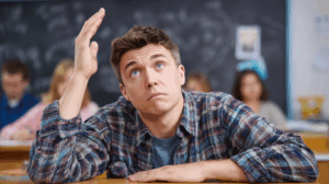 A person in a classroom, sitting at a desk with a puzzled look, raising their hand