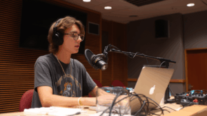 A student at a desk, speaking into a microphone and typing on a laptop. 