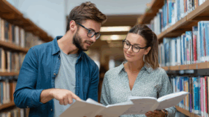 Two young professionals in a library discussing English grammar together, one pointing to a workbook with question forms.